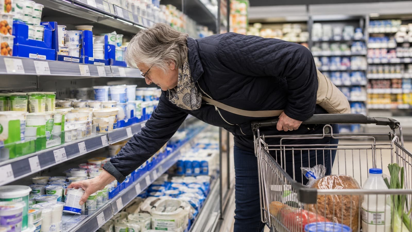 Rentnerin mit fast leerem Einkaufswagen greift im Discounter ins untere Kühlregal