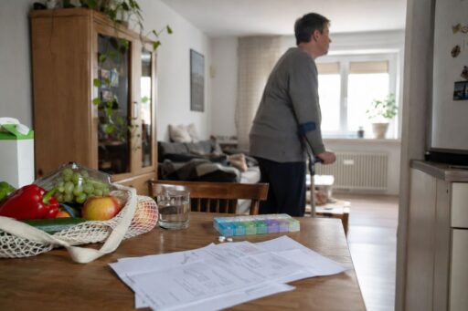 Person mit Gehhilfe in einer Wohnung, im Vordergrund liegen Unterlagen, Medikamente und Lebensmittel auf einem Tisch.