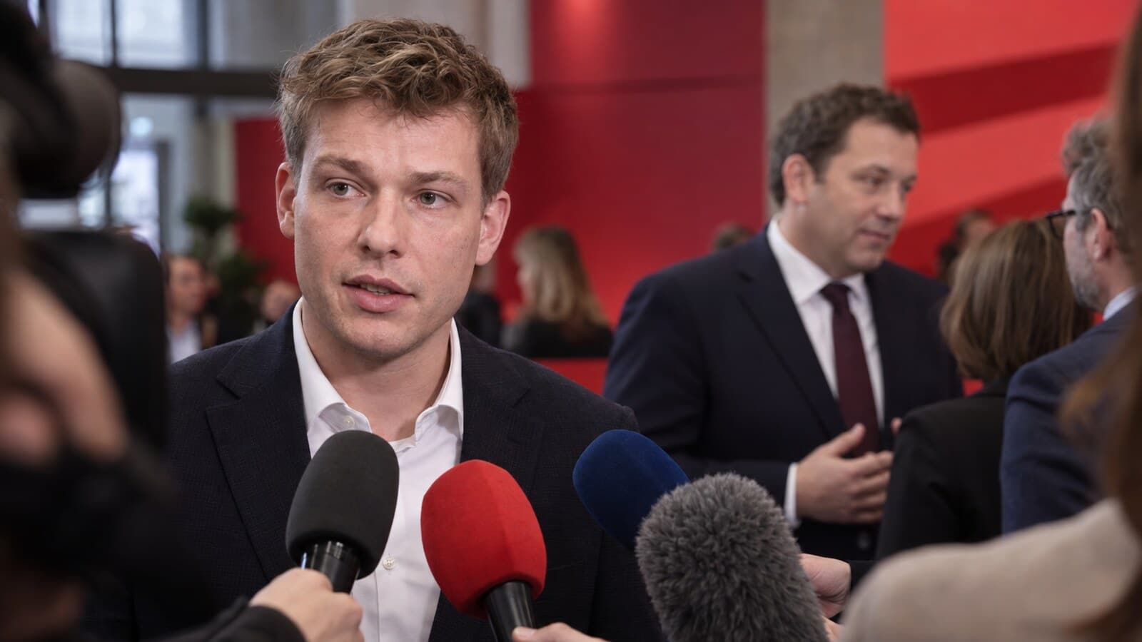 Tim Klüssendorf spricht vor Reportermikrofonen, während Lars Klingbeil im Hintergrund einer SPD-Kulisse steht.