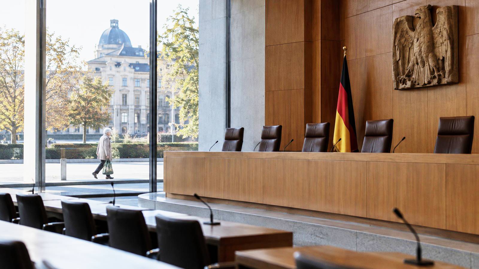 Leerer Gerichtssaal mit Richterbank, Bundesadler und Deutschlandflagge; draußen geht eine ältere Frau mit Einkaufstasche und Brief vorbei.