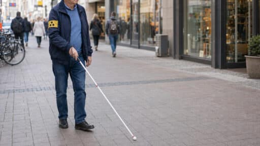 Sehbehinderter Mann mit gelber Armbinde führt seinen weißen Langstock durch eine belebte Fußgängerzone.