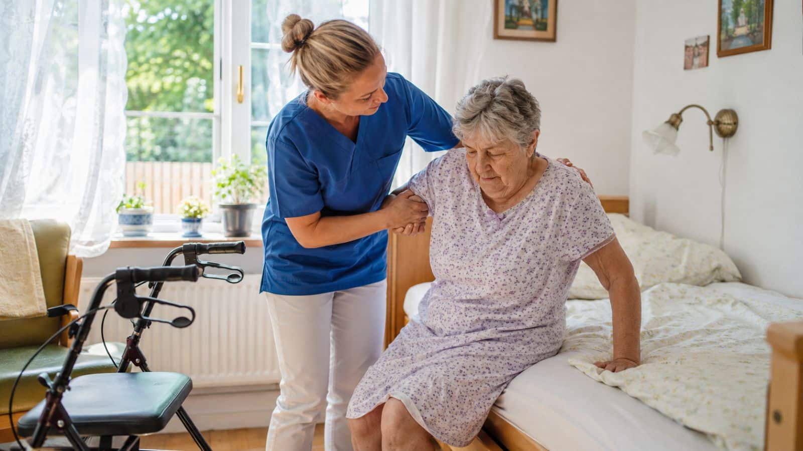 Pflegefachkraft in blauer Arbeitskleidung hilft einer pflegebedürftigen älteren Frau beim Aufstehen vom Bett in einer häuslichen Pflegesituation.