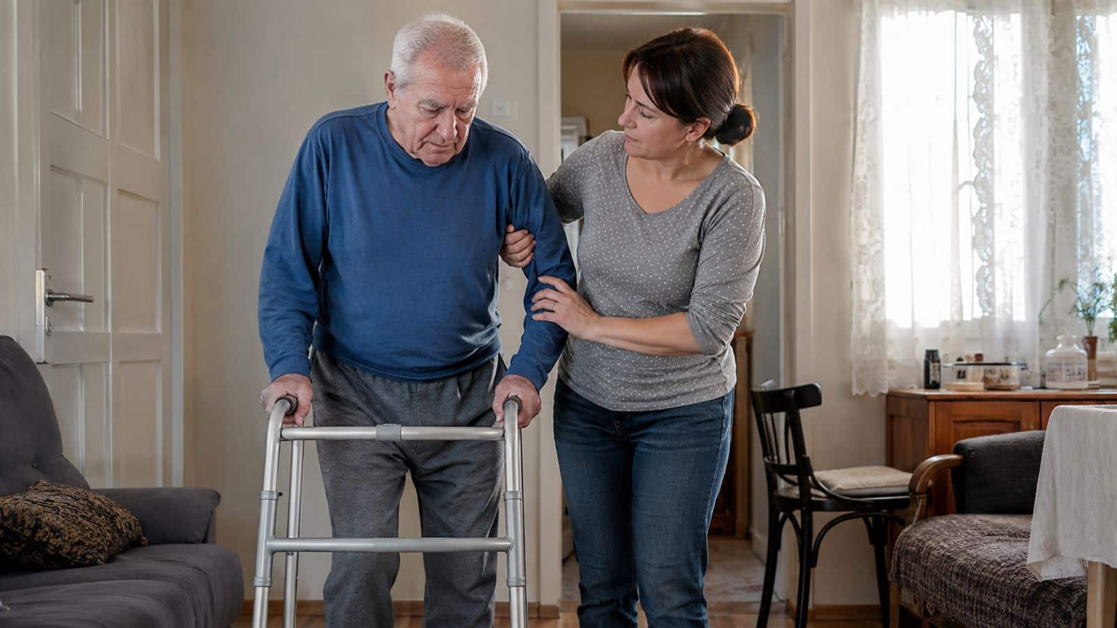 Tochter unterstützt ihren pflegebedürftigen Vater beim Gehen mit einem Rollator in der Wohnung.