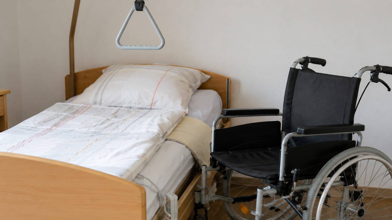 Leeres Pflegebett mit Aufrichter neben einem Rollstuhl