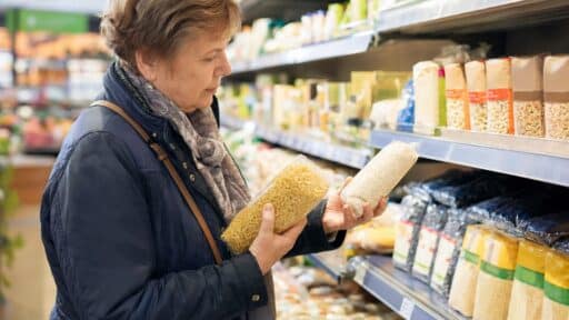 Beim Einkaufen vergleicht eine Kundin im Rentenalter die Preise zweier Grundnahrungsmittel im Supermarktregal.