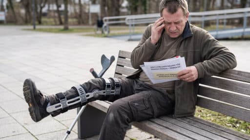 Mann mit Beinschiene und Gehstütze sitzt nachdenklich auf einer Parkbank und liest Dokumente im Freien