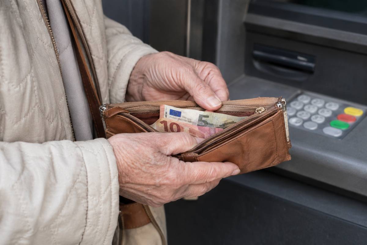 Rentner steht mit offener Geldbörse und wenigen Geldscheinen darin vor einem Geldautomaten
